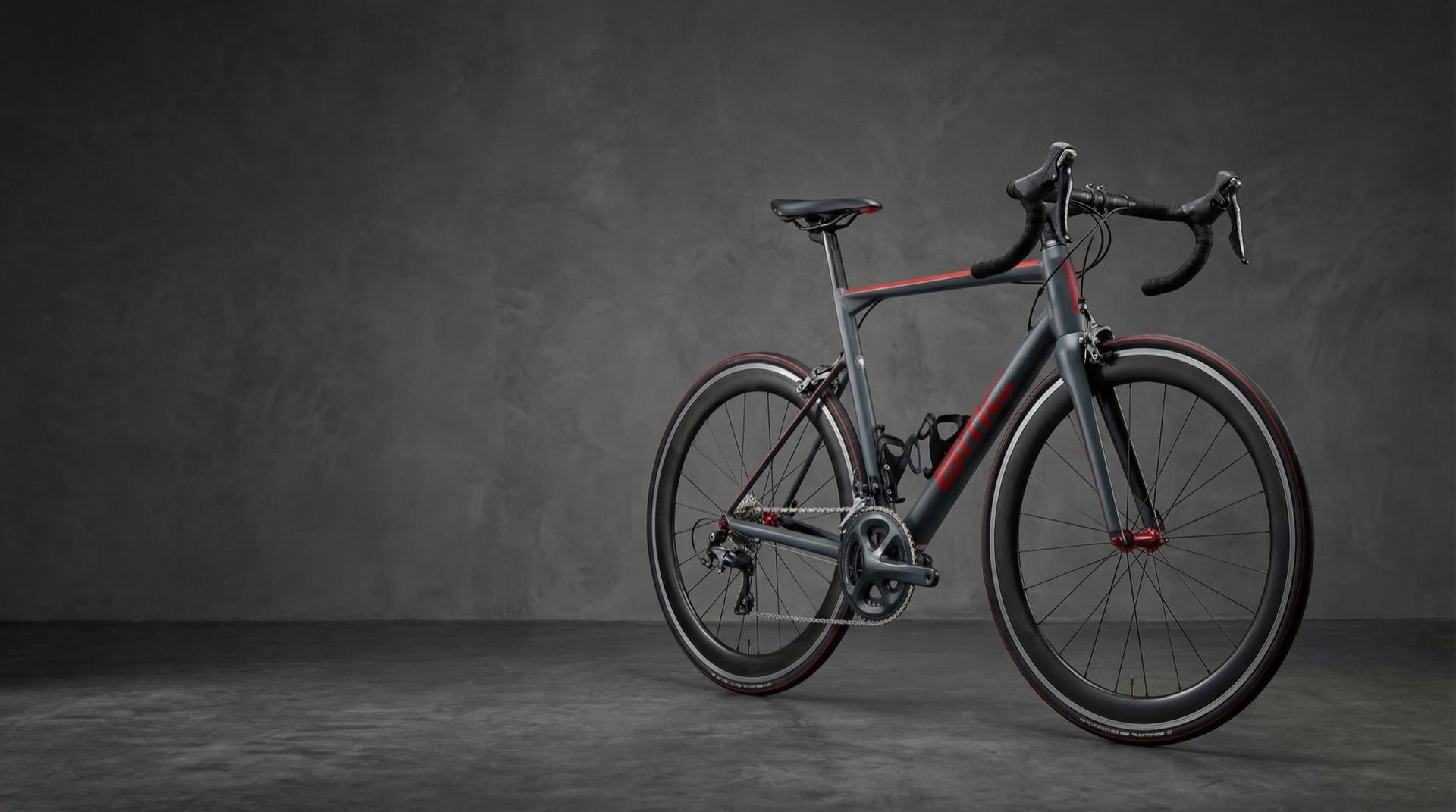 Grijze BMC racefiets met rode accenten en carbon wielen, studio met donkergrijze achtergrond